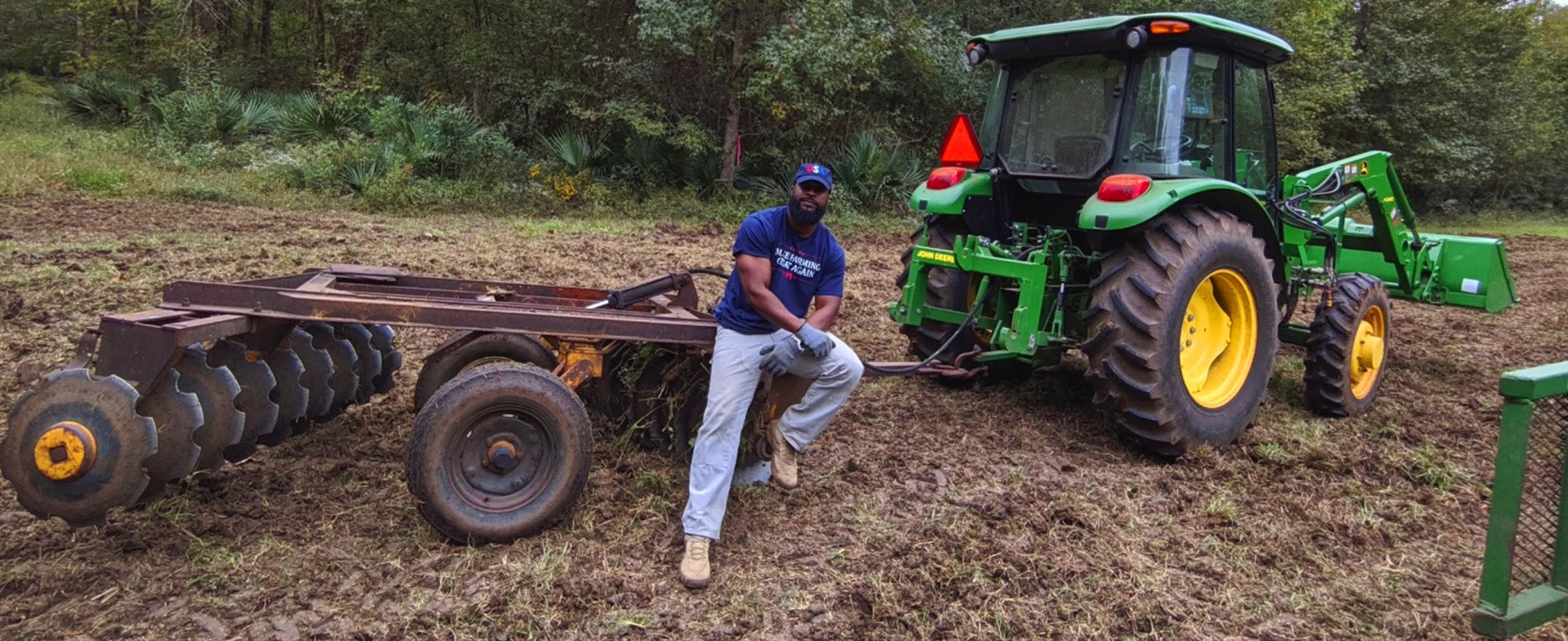 Christopher Joe, AL, wearing a blue t-shirt that says “Make Farming Great Again” and light blue jeans leaning on tractor equipment.